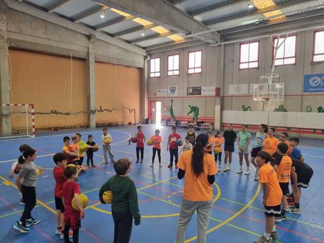 Xiquets i xiquetes amb pilotes atenent les explicacions d'una monitora en el poliesportiu