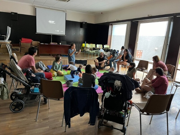 Famílies atenent les explicacions en una activitat APPI en el Centre Social Zona Nord mentre els xiquets xicotets juguen sobre una catifa